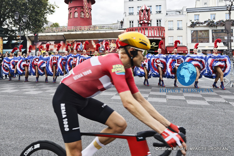 Tour de France riders race past the Moulin Rouge as dancers perform outside the iconic Paris cabaret.