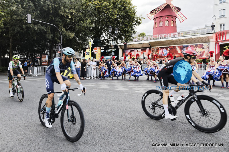 Tour de France cyclists ride past Moulin Rouge dancers performing outside the famous Paris cabaret.